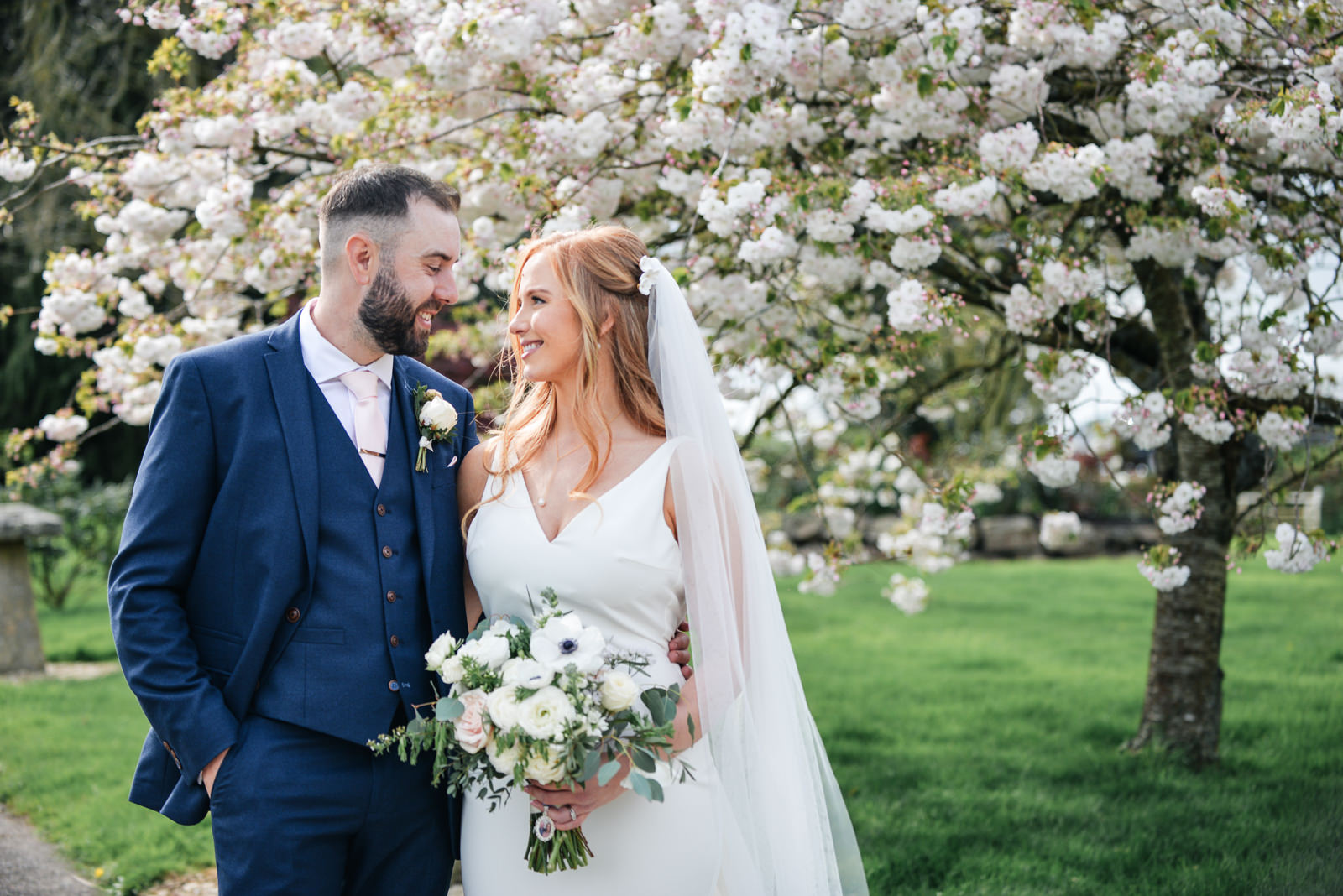 Bride and groom in front of a spring blossom tree smiling at each other taken at Upton Barn Collumpton